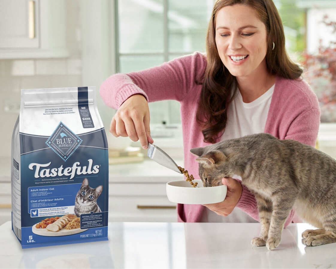 A woman happily serves food to her dog in the kitchen with a bag of premium kibble nearby.
