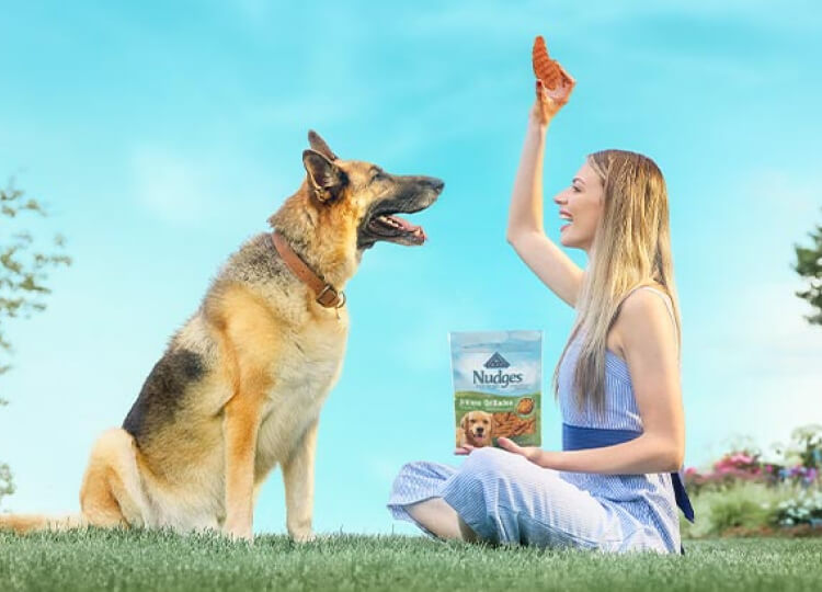 A woman is holding a treat for her German Shepherd. A bag of BLUE Nudges is in her other hand and they’re sitting on a lawn.