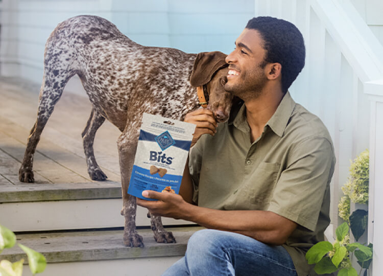 A man is feeding his dog a treat. A bag of BLUE Bits dog treats is in his other hand. They’re sitting on the steps of a porch.