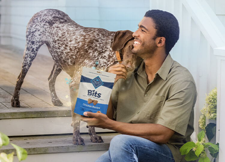 A man sits on the porch steps, smiling as he holds a bag of Blue Buffalo dog food. His cheerful dog approaches from the side, ready for a tasty treat.