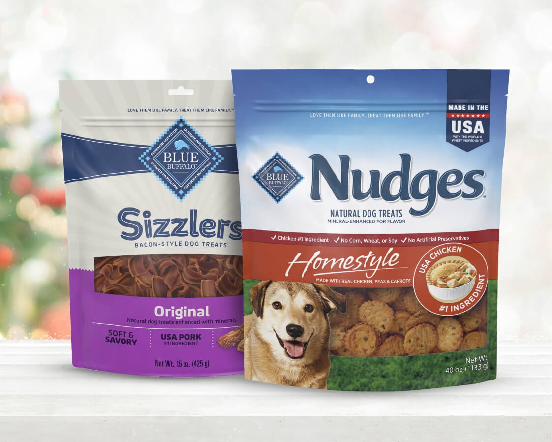 Two bags of Sizzlers Original and Nudges Homestyle dog treats rest on a festive red surface.