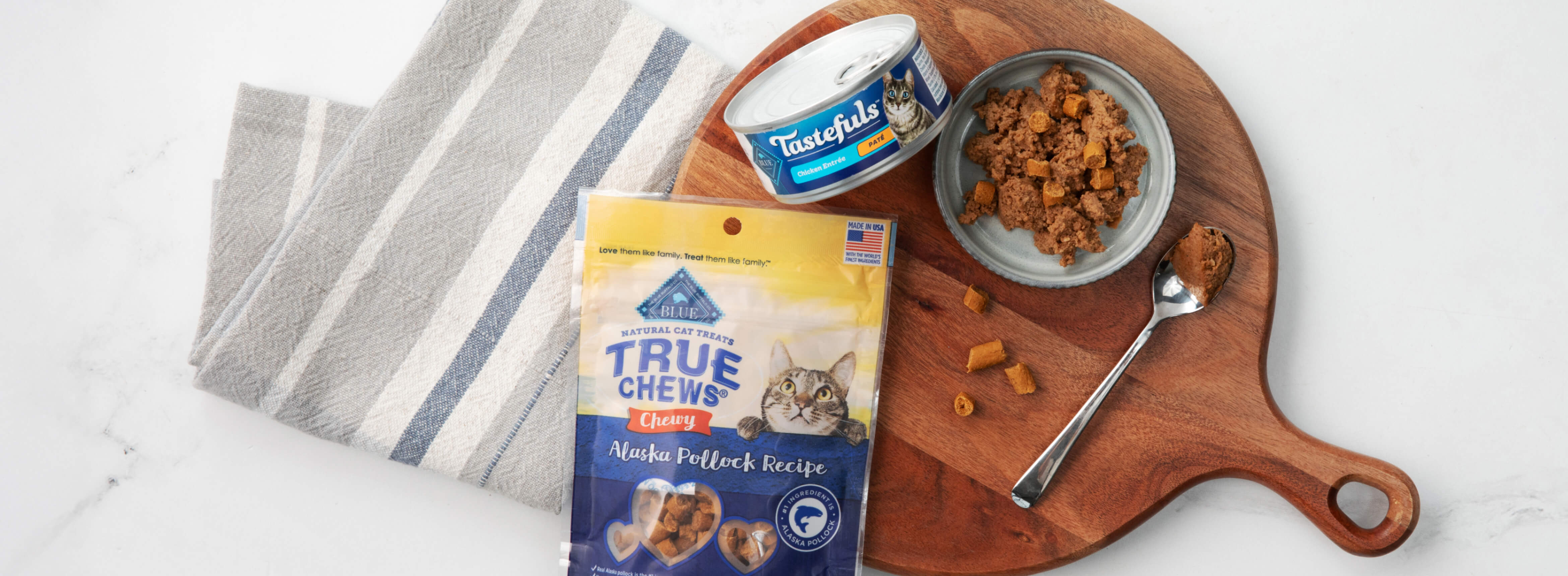 Bag of Blue Buffalo True Chews cat treats, a can of Tastefuls cat food, and a serving on a wooden board with a fork, next to a striped cloth.