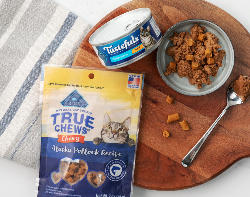 Bag of Blue Buffalo True Chews cat treats, a can of Tastefuls cat food, and a serving on a wooden board with a fork, next to a striped cloth.