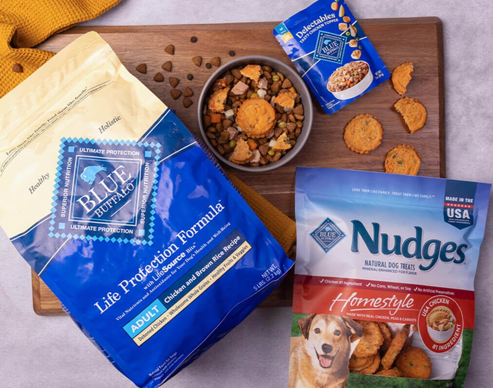 A dog-friendly meal sits in a bowl on a cutting board, surrounded by packages of BLUE Life Protection Formula, BLUE Delectables, and BLUE Nudges.