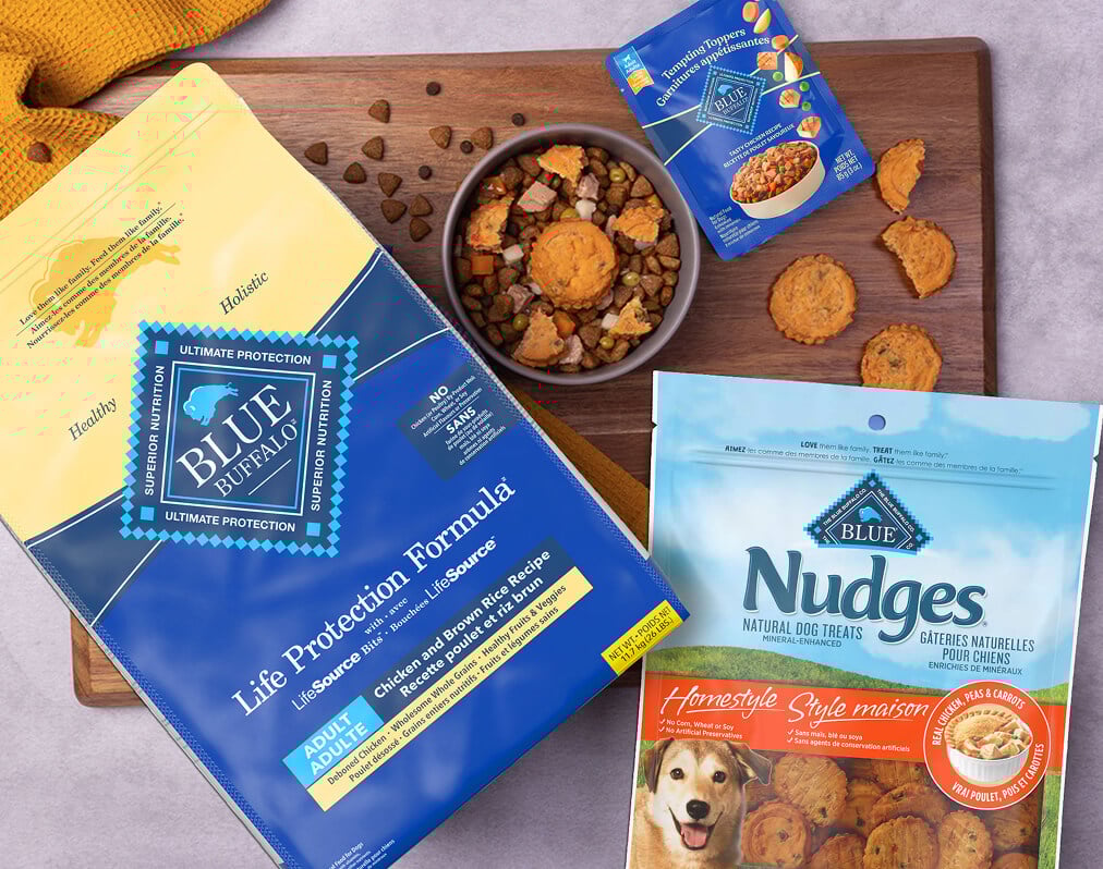 A dog-friendly meal sits in a bowl on a cutting board, surrounded by packages of BLUE Life Protection Formula, BLUE Delectables, and BLUE Nudges.
