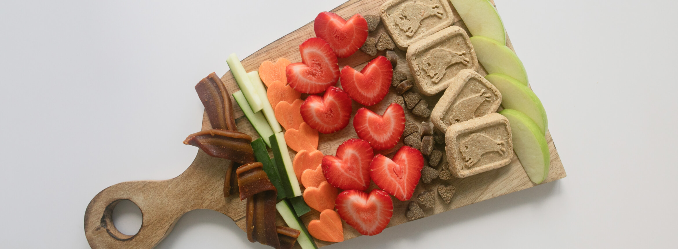 A wooden board with an assortment of dog treats and fruits and vegetables arranged in a visually appealing way.  The treats appear to be Blue Buffalo brand.  The fruits and vegetables include strawberries, apples, carrots, and cucumbers.